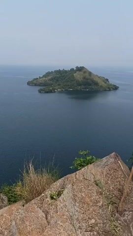 From a rocky cliffside vantage point, the video offers a view of a large, calm body of water, likely a lake or sea, with a small, lush island in the distance. The island is covered in dense vegetation, mostly trees, with a slightly elevated terrain. The water is a deep blue color, stretching to the horizon. The foreground is dominated by the rocky cliff, with sporadic greenery sprouting between the stones. The sky is a pale, hazy blue, suggesting a clear day with some atmospheric diffusion. The camera pans slowly from left to right to reveal more of the body of water.
