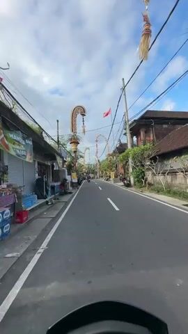 The video shows a POV of a person riding a scooter down a street. The street is paved and has white lines marking the lanes. On both sides of the street are buildings, some with traditional architecture and ornate decorations. Several scooters are parked along the sides of the road. Overhead, there are numerous electrical wires. Archways decorated with colorful ornaments and greenery span across the street at intervals. The sky is cloudy with patches of blue. A red flag is visible on a pole in the distance. The general atmosphere appears to be a village or town, possibly in Southeast Asia, given the architecture and decorations.