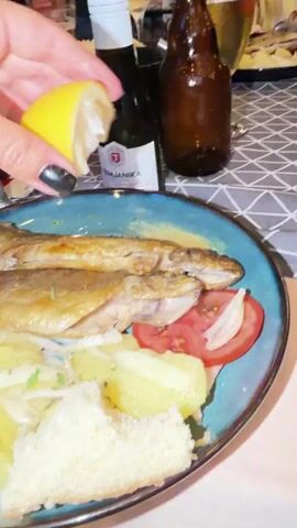 The media file shows a close-up of a person's hand squeezing lemon juice onto a plate of food. The plate contains two whole cooked fish, roasted potatoes, sliced tomatoes, and thinly sliced onions, garnished with chopped parsley. A piece of bread is also on the plate. In the background, there are bottles of drinks and a patterned tablecloth, suggesting a restaurant setting. The lighting appears to be artificial, indicating an indoor scene, possibly in the evening. The overall mood is relaxed and focused on the act of preparing the meal.

Visible text on a label in the background reads "TEMJANIKA". While not clearly legible in full, the word "ANIKA" is visible on a menu or label in the background. Given the location context of Travnik, Bosnia and Herzegovina, this is likely a local restaurant.