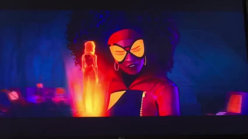 The media file is a short video clip featuring an animated character, presumably a superhero, with dark, curly hair and a red mask with yellow accents covering her eyes. She is wearing a red outfit and appears to be in a dimly lit, futuristic or laboratory-like setting. The background is a mix of dark blues and purples, with some glowing elements and what look like computer screens or consoles. To her left, there is a holographic projection of a person, glowing with an orange and yellow light, that seems to be emanating from her hand. The character's expression is serious and determined. The overall mood is intense and possibly suspenseful. There is no visible text within the clip. The location context of Vozrozhdeniye, Russia, does not appear to be visually represented in the clip itself.