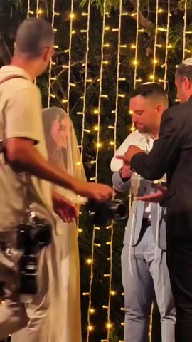 A wedding ceremony is taking place outdoors at night, with a backdrop of numerous small, warm-toned lights strung vertically. A bride, wearing a white wedding dress and veil, stands on the left. Facing her is a groom in a light blue suit, who is being draped with a tallit (Jewish prayer shawl) by another man. The groom then lifts the veil from the bride's face and they share a tender moment, leaning in as if for a kiss. The groom adjusts the tallit.