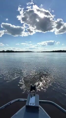 This media file captures a serene scene on a wide body of water, likely a river, under a bright, partly cloudy sky. The sun is visible through a large, fluffy cloud, casting shimmering reflections on the water's surface. The foreground shows a portion of a boat, including a metal fitting with a pulley, indicating the perspective is from a vessel in motion. In the distance, a faint outline of a bridge can be seen spanning the waterway, and the shores are lined with trees, suggesting a natural, possibly rural, environment.

The activity taking place is a boat ride, evidenced by the ripples and wake on the water and the perspective from the boat. The time of day appears to be late morning or early afternoon, given the bright sunlight and the angle of the sun. The weather is fair and pleasant, with a clear blue sky dotted with white clouds. The location cues, along with the provided context of Sosnovka, Russia, suggest this is a scene on a Russian river. There are no people explicitly visible, but the presence of a sailboat in one frame indicates recreational activity on the water. The overall mood is peaceful and tranquil, characterized by the calm water, the expansive sky, and the gentle movement of the boat. No text is visible in the media file.