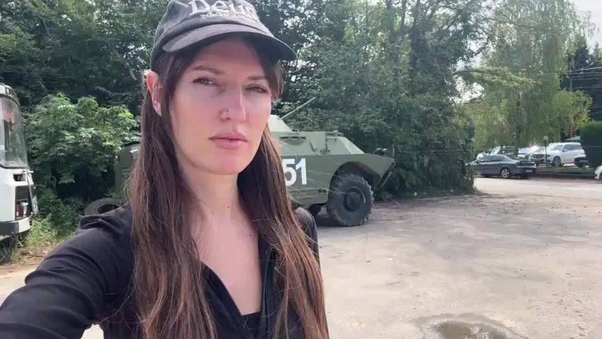 A medium shot shows a woman wearing a black shirt and a black baseball cap. She is looking at the camera with a neutral expression. Behind her, there is a military vehicle with the number 351 on its side, and a white bus on the left. The background consists of trees and a dirt road. The woman's face is partially obscured by her hair. The video is shot outdoors on a sunny day.