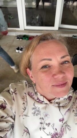 The video shows a woman taking a selfie. Initially, the camera is tilted downwards, showing her floral-patterned blouse and black pants. She then raises the camera to reveal her face, speaking directly to the camera. The background shows a cluttered entryway. She continues to move the camera around, showing a table set for a meal with several people seated around it. The table is covered in dishes, food, and drinks. In the background, a balcony overlooks a scenic view of trees, buildings, and a distant body of water.