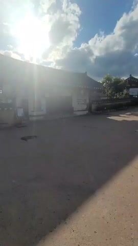 A video begins with a bright sun casting rays across a dirt path and traditional buildings. The buildings have dark, tiled roofs with curved eaves. There are some plants and trees around the buildings. A stone sculpture of several figures and a tall, thin object stands on the right side of the path. The path continues into the distance, lined with more traditional buildings and some decorative flags. The sky is blue with fluffy white clouds. The camera pans slowly to the right, revealing more of the scene.