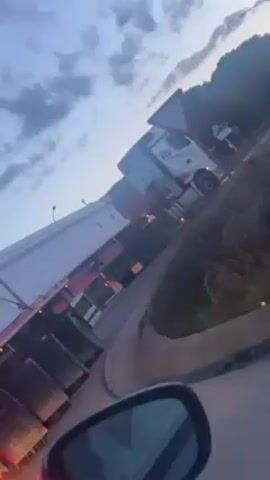 The video shows a white semi-truck and trailer struggling to navigate a sharp turn on a road. The truck is partially blocking the lane. A yield sign is visible near the truck. The footage is taken from a car stopped behind the truck. The sky is cloudy and it appears to be dusk.
