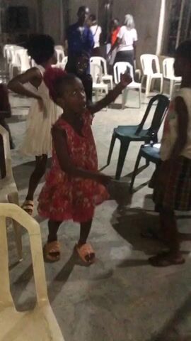 The video captures an indoor scene, likely a community hall or a large room in Ukwu-Abwa, Nigeria. The primary subjects are several children energetically dancing, with adults visible in the background.

In the foreground, a girl wearing a red floral dress and light-colored sandals dances with enthusiasm. To her left, another girl in a white dress also moves lively. On the right, a third child in a plaid skirt and light-colored top is seen dancing. The children's movements are uninhibited and joyful, suggesting a celebratory or communal atmosphere.

Behind the dancing children, several adults stand and observe, some appearing to also move to the rhythm. The setting includes numerous white plastic chairs arranged in rows, with a few dark chairs also present. The floor appears to be concrete. The lighting is dim, indicating that the activity is taking place in the evening or under artificial light. There is no visible text in the video. The overall impression is a vibrant gathering where children are expressing joy through dance.