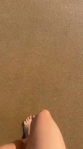 A first-person perspective video shows feet walking on a wet sandy beach. The camera is angled downwards, focusing on the legs and feet as they move through shallow water. The water washes over the feet and ankles, creating foam and ripples. The background shows the ocean with small waves and a distant shoreline with trees. The sunlight casts a shadow of the legs and feet on the sand.