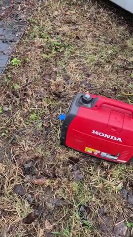 The video shows an outdoor scene featuring a variety of equipment positioned on a flat, possibly metal, surface covered with moisture and some debris. There is a black, hard case labeled “Powercase,” a yellow toolbox, and a red and black generator. A metal chain is draped on the surface, and a black cable is coiled nearby. The camera then shifts to reveal an open rectangular pit below the surface. Inside the pit, a rusty pipe with fittings is visible, and the bottom of the pit appears to be filled with murky water. The environment surrounding the equipment and the pit consists of grass, fallen leaves, and bare ground.