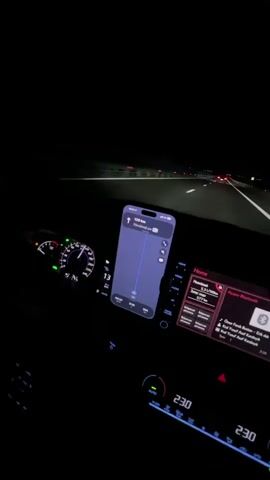 The video captures the interior of a car at night, as seen from the passenger's perspective. The dashboard lights are illuminated, showing a speedometer with a needle indicating around 130 km/h. A smartphone mounted on the dashboard displays a navigation app with a route and distance remaining. The car's infotainment system is also visible, showing music playback information. The road ahead is dark, with the headlights of other vehicles visible in the distance. The overall impression is of a nighttime drive on a highway.