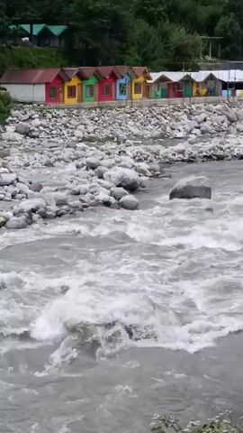 The video displays a fast-flowing, murky river with white rapids. The riverbed is composed of large, grey stones. On the left bank, a row of small, brightly colored cabins with red, yellow, green, and blue roofs are visible. Behind the cabins, lush green trees and foliage are present. The overall scene depicts a natural landscape with man-made structures situated near a powerful river.