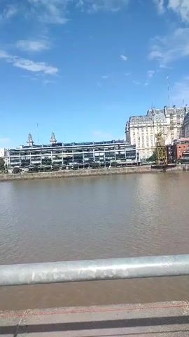 The video shows a cityscape viewed from across a river. The camera pans from right to left, revealing buildings and a large, white sailing ship docked in the distance. The river is a murky brown color. The foreground shows a grey metal railing. The sky is blue with scattered white clouds. The overall impression is a bright, sunny day with a clear view of the city.