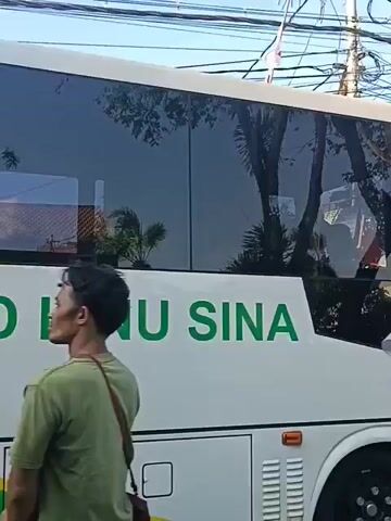 The video shows a white ambulance with red text reading "AMBULANCE TRANSPORT". A green cross symbol is visible on the side of the vehicle. Two women in hijabs are standing near the front of the ambulance. One woman is wearing a light purple hijab, and the other is wearing a black hijab. Behind them, a man is taking a photo with his phone. Further down the road, several people are standing near the back of the ambulance, where the doors are open. The environment appears to be an outdoor street scene with buildings and overhead electrical wires.