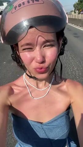 Here is a description of the video.

The video shows a fair-skinned young woman with rosy cheeks riding on what appears to be a motor scooter on a highway. She is wearing a light blue, strapless top and a white beaded necklace. She is also wearing a pink helmet with the name "Polly" written on it, and goggles, that are up on her helmet. The background reveals a sunny day, with a clear blue sky overhead, and highway traffic, including a truck, passing by. The young woman makes silly faces, then waves to the camera. A glimpse of a forearm tattoo is visible when she raises her arm.