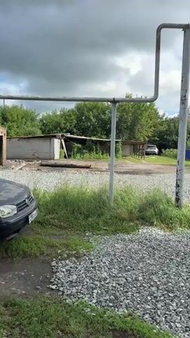 A car is parked in front of some dilapidated structures. One structure appears to be a garage or shed with a corrugated metal roof that is partially collapsed. Another structure is made of brick and has a blackened facade, suggesting it may have been affected by fire. A car is parked in the foreground and another vehicle, possibly a white SUV, is visible in the distance. The ground is covered in gravel and patches of grass. Overhead, a metal pipe structure runs across the frame. The sky is cloudy.