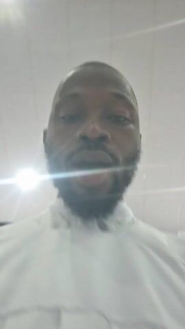A close-up video shows a man with a beard wearing a white garment. He is looking directly at the camera and raising his right index finger upward. The background is a white ceiling with bright lights.