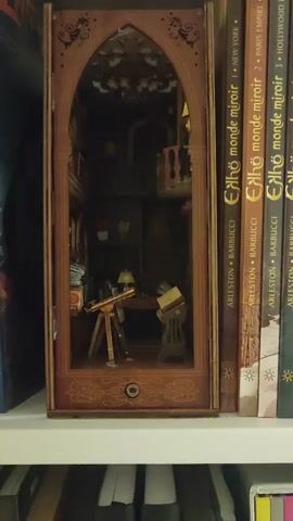 The video shows a close-up view of a miniature diorama depicting a study or library. The diorama is housed within a wooden frame with an arched opening, resembling a doorway or window. Inside, the scene is dimly lit and features a desk with a lamp and an open book on a stand. To the left, there is a telescope on a tripod and a stack of books. The background includes shelves and a balcony. The diorama is positioned on a shelf next to several books.
