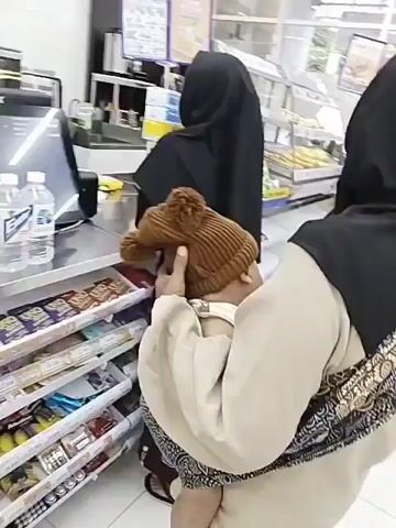 The video shows a dimly lit convenience store with shelves stocked with merchandise. In the foreground, a person wearing a tan sweater and a patterned baby carrier is holding a baby wrapped in a tan beanie. The baby is facing the camera. Behind them, a woman in a black hijab and patterned skirt stands at a checkout counter, with her back to the camera. To the right, another person in a gray sweatshirt and black pants stands looking at the checkout counter. The overall environment appears to be a typical retail setting.