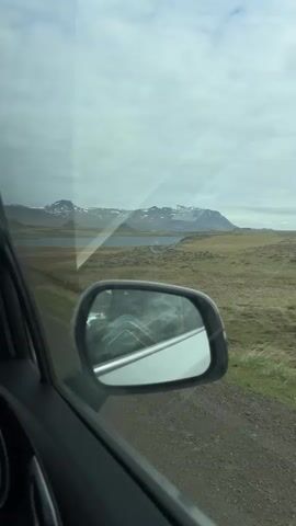 The video shows a view from inside a moving car, looking out the passenger window. The car is traveling on a gravel road through a rugged, expansive landscape, likely in Iceland as indicated by the provided context (Borgarnes). In the foreground, the dark grey frame of the car door and the side mirror are visible, reflecting a distorted view of the car's exterior and the road.

Beyond the car, the scenery features vast stretches of green and brown, low-lying vegetation, characteristic of Icelandic terrain. In the middle distance, a calm body of water (possibly a lake or fjord) reflects the overcast sky. Further in the background, majestic snow-capped mountains rise under a cloudy sky, their peaks partially obscured by the clouds. The overall weather appears overcast but clear, with good visibility of the distant mountains. The time of day seems to be daytime, with soft, diffused light. There is no visible text in the video. The scene evokes a sense of quiet travel through a starkly beautiful natural environment.