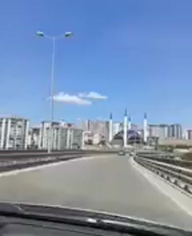 This media file captures a video from a vehicle, likely a car, as it travels along a highway. The primary subject is a modern mosque with a large, distinctive blue domed roof and four tall, slender white minarets topped with black spires. The mosque is situated in the background, appearing to be under construction or recently completed, as some scaffolding and unfinished elements are visible.

Surrounding the mosque are several tall, modern residential buildings, suggesting an urban or suburban setting. The highway itself is the foreground, with clear lane markings and guardrails visible. The sky is a vibrant, clear blue with a few wispy white clouds, indicating fair weather, likely during the daytime. The sunlight is bright, casting shadows and illuminating the scene.

The perspective is from the driver's or front passenger's seat, with the dashboard visible in the lower part of the frame. The camera is angled slightly upwards, focusing on the mosque and the cityscape. The movement of the vehicle creates a sense of journey and observation. No people are clearly discernible within the frame, though it is possible they are present inside buildings or other vehicles. No text is visible in the media file. The overall impression is one of observing a significant architectural landmark in a developing urban landscape under a bright, clear sky. The location context provided places this scene in Kırıkkale, Turkey.