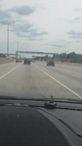 The video captures the view from the dashboard of a car driving on an open road tollway. The car is moving forward on a multi-lane highway under a cloudy sky. The dashboard has a black, textured surface with a small, black circular object placed in the center. In the distance, there are several cars on the highway. A large overhead structure with signs and equipment, typical of an open road toll system, is visible. The signs on the overhead structure are mostly unreadable but appear to indicate toll information. The environment appears to be a typical highway setting with some trees and vegetation visible on the sides of the road in the background. The perspective is from the driver's seat, looking straight ahead through the windshield.