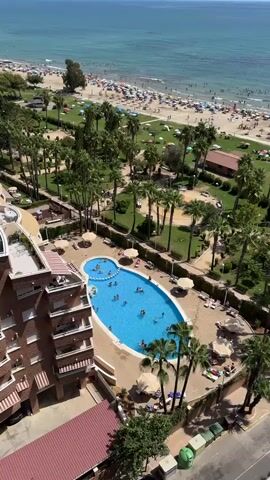 The video shows an aerial view of a resort or hotel property next to a beach. In the foreground, there is a multi-story building with balconies. Adjacent to the building is a kidney-shaped swimming pool filled with blue water and several people swimming and playing. Around the pool, there are lounge chairs and umbrellas, some occupied by people. A paved area surrounds the pool, and beyond that, a park-like area with numerous palm trees and grassy patches. Further in the background, a wide sandy beach is visible, crowded with sunbathers and umbrellas. The sea is in the distance, with a clear blue horizon line. The overall scene depicts a sunny, populated vacation destination.