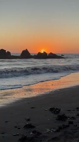 This is a vertical video clip of a beach at sunset. The sun is low in the sky, casting an orange glow and reflecting on the wet sand. Waves are crashing onto the shore, and rocks are visible in the ocean. The sand is dark and wet, with some seaweed scattered on it. The sky is clear and transitions from orange at the horizon to a lighter yellow and then a pale blue.