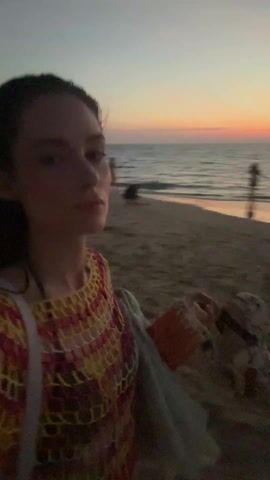 A young woman stands on a beach at sunset, holding the camera at arm's length and filming herself. She is wearing a colorful, crocheted top and has wet hair. In the background, the sky is a mix of orange, pink, and blue as the sun sets over the ocean.  There are a few other people on the beach, some near the water and others sitting on the sand. As the video progresses, the light dims and the sky turns a darker blue, transitioning from sunset to twilight.
