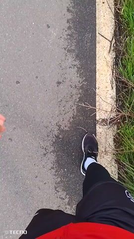 A person is walking on a paved path. The camera appears to be positioned looking down at the person’s legs. The person is wearing black athletic pants, a red top, white socks, and dark-colored athletic shoes with white accents. The path is bordered by a white concrete edge, with grass and foliage visible beyond the edge on one side. The surface of the path appears slightly damp. The video is taken from a first-person perspective, looking down at the legs of the person walking. The logo “TECNO” is visible in the bottom right corner of the frame. Sounds of traffic can be heard in the background.