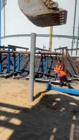 A construction site with a large white tank in the background. In the foreground, an excavator bucket is positioned above a vertical pipe that is partially buried in sand. Two construction workers, wearing orange vests and hard hats, are working near the base of the pipe. One worker is standing, and the other is kneeling. Blue and brown metal structures are visible behind the workers, and black sheeting covers the ground. The scene appears to be under a clear blue sky.