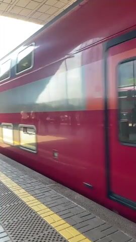The video shows a red and gray double-decker train moving past a train platform. The train is traveling to the right and people can be seen sitting inside it through the windows. The platform is made of brick tiles and has yellow safety lines painted on the edge. There are several people standing on the platform, some with luggage, waiting to board. The sky is bright, and the sun is shining on the train.