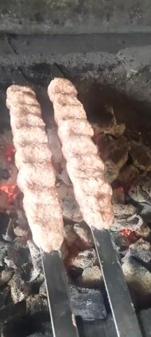 The video shows several skewers of ground meat being grilled over hot coals. The meat is shaped into elongated, ribbed cylinders on metal skewers. Red embers are visible beneath the skewers, indicating intense heat. Smoke rises from the grilling meat, and some of the skewers appear to be slightly charred. The overall scene depicts the process of cooking kebabs on an outdoor grill.