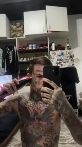 Here's a detailed description of the video:

A shirtless man with extensive tattoos on his chest, arms, neck, and face is taking a selfie video while shaving the side of his head. He is using an electric trimmer/razor in his right hand, which is pink and black. He holds his phone in his left hand, using it to record himself in a mirror. The phone is black, and he's looking at the screen as he shaves.

The background is a bedroom interior. Behind him is a white wardrobe with open shelving. Various items are visible on the shelves, including clothes, shoes, and decorative objects. Some items on the top shelf appear to be small figures or toys.

The man's hair on top of his head is styled and longer than the area he is shaving. He has stretched earlobes (gauges). The room is generally well-lit.