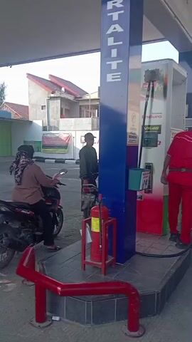 A male gas station attendant wearing a red uniform is dispensing fuel from a gas pump into a motorcycle. A woman wearing a hijab is sitting on the motorcycle being refueled. Another person, appearing to be male, stands near a motorcycle to the left of the gas pump. A fire extinguisher is visible on the left. The scene appears to be outdoors during the day, with buildings visible in the background.