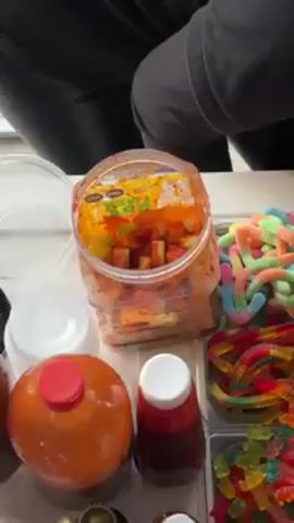 A high angle view shows a container of diced fruit, including watermelon and melon, covered in red chili powder. The container is sitting on a white surface with other containers of colorful gummy worms, chili sauce, and lids. A person wearing black appears to be picking at the contents of the container.