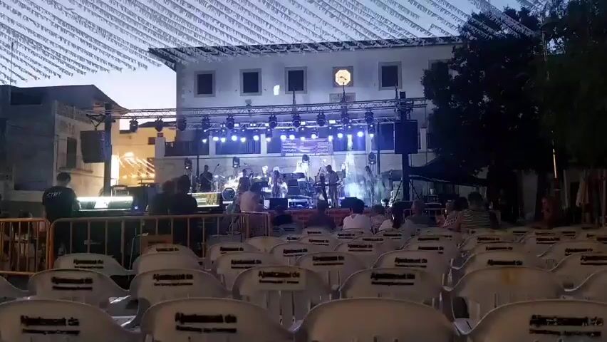 The video shows an outdoor concert setup at dusk. In the foreground, rows of empty white plastic chairs are visible, some with a logo that appears to say "Ajuntament de". Beyond the chairs, a stage is set up with a band performing. The stage is lit with blue and white lights. Several band members are visible on stage, appearing to play instruments. The backdrop to the stage is a building with windows and a clock. Strings of small flags or lights are strung across the sky above the stage and the building. People are seated in chairs near the stage, facing the performers. Some individuals are standing behind barriers in front of the stage. The overall environment appears to be a town square or plaza.