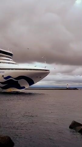 The video shows the Sapphire Princess cruise ship sailing on the water. The ship is white with blue wave-like designs and the words "SAPPHIRE PRINCESS" and "PRINCESS CRUISES" printed on its side. There are several decks visible, with rows of windows and balconies. Lifeboats are also visible along the side of the ship. The water is dark and appears somewhat choppy, and the sky is cloudy and overcast. In the foreground, there are some rocks and what appears to be a portion of a dock with yellow and black stripes. The ship is moving from left to right across the frame, with the front of the ship visible on the left side of the initial frames and the ship progressively moving further into the frame.