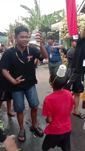 A group of children and young adults are gathered outdoors, participating in what appears to be a game. Several individuals, both children and older participants, have plastic cups balanced on their heads. A red and white flag, likely a national flag, is visible hanging in the background, suggesting a festive occasion. Some participants are holding thin strings or threads suspended from above, which they are manipulating. One boy in a red shirt and black shorts is facing away from the camera, with a cup on his head. Another boy in a yellow shirt and beige shorts is also facing away, with a cup on his head. Several older youths are actively engaged in the game, some holding strings and appearing to concentrate on balancing the cups. The environment is somewhat rural or suburban, with buildings and trees in the background. The ground is wet in places, possibly from recent rain or as part of the game. The overall atmosphere seems to be one of lively, outdoor recreation and competition.