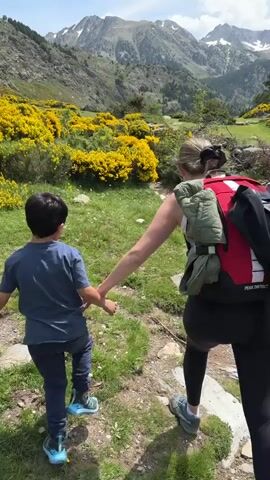 A child and a woman walk away from the camera on a dirt path in a mountainous region with green and yellow plants and bushes. The child on the left wears a blue shirt and pants and holds the woman's left hand. The woman on the right wears a black top and pants, a red backpack, and a white bag hanging from her left hand. Mountains with snow patches are visible in the background under a blue and cloudy sky.