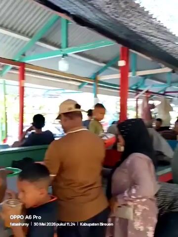 The video shows a bustling outdoor market or gathering space. Several people are visible, including men, women, and children. Some women are wearing hijabs. People are standing around and walking through the area. Tables and benches are positioned throughout the space, with various items displayed on them. A man is holding an inflatable donut-shaped swimming ring. The area is covered by a roof supported by green and red posts. The camera moves slightly, capturing different areas of the crowd. Text at the bottom of the video indicates it was taken on an OPPO A6 Pro on May 30, 2026, at 11:24 in Kabupaten Bireuen.