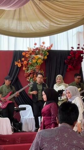 The video shows a wedding reception. A band is performing on a small stage with a red and black backdrop decorated with orange and red flowers. Several guests are standing near the front of the stage, including women in hijabs and men in casual attire. The camera pans across the crowd, focusing on the band and the guests. At the end of the video, the camera briefly focuses on a large black fan in the foreground. The atmosphere seems celebratory, with music and people gathered together.
