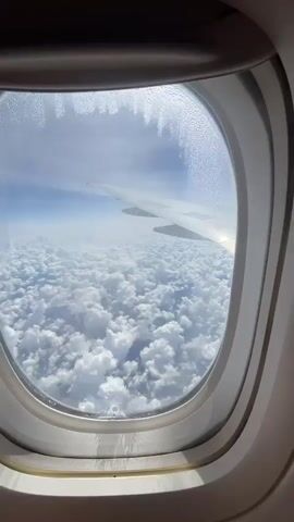 This video shows a view from an airplane window. The window frame is visible around the edges. Outside, a thick layer of white, fluffy clouds fills the lower part of the view. Above the clouds, the sky is a clear blue, with a hazy horizon. The wing of the airplane extends diagonally across the upper right portion of the frame. Sunlight reflects off the wing. There are water droplets condensed on the inside of the airplane window, forming a pattern along the top edge.