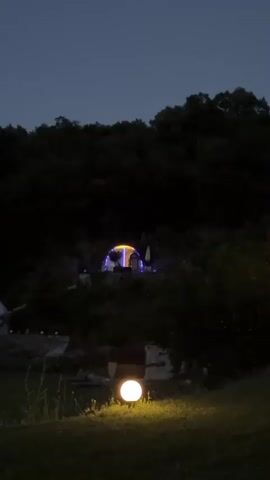The video begins with a wide shot of a dark, forested hillside at twilight. A round, illuminated orb is visible in the foreground, casting a soft yellow glow on the grass. In the mid-ground, nestled amongst the trees, is a dome-shaped structure or tent, illuminated with colorful lights, cycling through green, blue, yellow, and pink. The overall mood is serene and slightly mysterious, with the natural landscape contrasting with the artificial lighting of the dome. The camera remains stationary throughout the short clip.