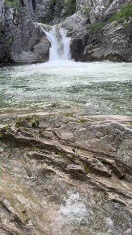 The video shows a small waterfall cascading into a rocky pool. The waterfall is surrounded by rocks and some vegetation. The water is clear and turbulent where it falls. The foreground consists of layered rocks with some moss growing on them, partially submerged in shallow water. The general environment seems to be a natural, outdoor setting, possibly a mountain stream or a secluded part of a river.