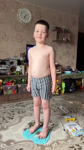A young boy stands on a patterned rug, wearing striped boxer shorts. He is positioned on two light blue orthopedic insoles, each with numerous raised nubs designed to stimulate the soles of his feet. The boy is shirtless and has short, dark hair. His facial expression is a wide, toothy grin, conveying happiness and playfulness.

The background suggests an indoor domestic setting, likely a child's bedroom or living area. A television is visible behind the boy, and shelves filled with toys and other items are present. A clock on the wall indicates the time. The room appears somewhat cluttered, with children's toys scattered on the floor and shelves, hinting at an active play environment. The lighting suggests it's daytime. There are no visible text elements in the frames, other than numbers on the clock.