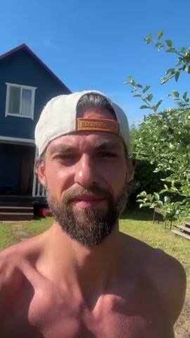 This is a selfie video of a shirtless man wearing a white baseball cap. He has a beard and is looking directly at the camera with a slight smile. The setting appears to be outdoors, possibly in a backyard, with a blue house and green trees in the background. The sun is shining brightly, indicating a clear day. There are some wooden benches and a grill visible in the background, suggesting a casual outdoor living space. The time of day appears to be midday, given the bright sunlight and clear blue sky. The man's relaxed expression and the sunny outdoor setting suggest a leisurely or casual moment. There is no visible text in the media file.