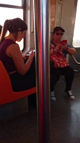 The video captures a scene inside a subway car. Several passengers are visible, some seated and others standing. A woman in a black tank top and jeans stands in the middle of the car, singing into a microphone. Another passenger, seated, is using a cell phone. The subway car has stainless steel handrails and doors, and the floor is gray. The lighting inside the car is bright. The video is taken from a seated passenger's perspective, showing a portion of their lap.