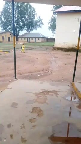 A person in a yellow uniform walks across a muddy, wet courtyard. The person is carrying a black backpack. There are buildings in the background. The sky appears overcast.