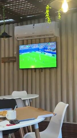 This is a video of a television screen displaying a soccer match. The television is mounted on a wall with vertical textured paneling. An air conditioning unit is installed above the television. In the foreground, there are white tables and chairs. One table has a white mug, a plate with food, and a book on it. The scene appears to be in a cafe or restaurant with an outdoor or semi-outdoor seating area, indicated by the overhead structure with hanging plants and lamps. The lighting suggests it is daytime. The soccer game on the screen shows players in white and blue uniforms on a green field.