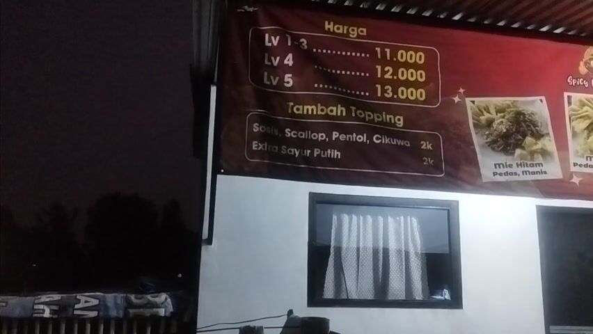 This media file captures a nighttime view of a food stall, likely a noodle shop, in Prajuritkulon, Indonesia. The primary focus is a large, red banner advertising menu items and prices.

The banner displays "Harga" (Price) with several "Lv" (Level) options: Lv 1-3, Lv 4, and Lv 5, with corresponding prices of 11.000, 12.000, and 13.000 (likely Indonesian Rupiah). Below this, there's a section for "Tambah Topping" (Add Topping), listing "Sosis, Scallop, Pentol, Cikuwa" for 2k, and "Extra Sayur Putih" (Extra White Vegetables) also for 2k.

To the right of the pricing, there are three images of food. The first is labeled "Mie Hitam Pedas, Manis" (Black Noodles Spicy, Sweet). The second is "Mie Putih Pedas, Asin, Gurih" (White Noodles Spicy, Salty, Savory). The third is "Pangsit Goreng Ayam" (Fried Chicken Wontons). A small logo with a cartoon character and the text "Spicy Noodles" is also visible. Further to the right, a menu section lists "Kopi Wedang Jahe, Wedang Kencur, Es Teh, Pop Ice, Nutrisari" (Coffee, Ginger Drink, Galangal Drink, Iced Tea, Pop Ice, Nutrisari). Contact information and a location "Jl. Prajuritkulon" are also present, along with a phone number and a website or social media handle.

The scene is dimly lit, indicating nighttime. A white building wall forms the backdrop for the banner. A black-framed window with white curtains is visible below the banner. To the right of the window is a dark doorway with a sign that reads "TUTUP" (Closed). There are some objects on a shelf or table to the left of the window, including what appears to be folded clothes. The overall impression is of a small, local food establishment operating after dark. No people are clearly visible in the frame. The weather appears to be clear, given the absence of any rain or strong wind effects.